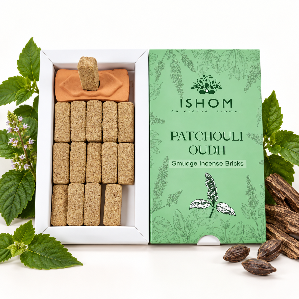 An open white box of Ishom Patchouli Oudh smudge incense bricks next to its green illustrated packaging box, styled with fresh green leaves, small flowers, and dried bark on a white background.