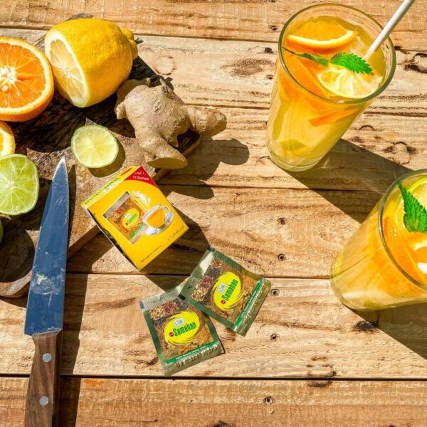 A tea product setup on a rustic wooden table with two glasses of orange tea garnished with lemon slices and mint, a box and two tea bags labeled Simla, ginger, lime, and orange slices, plus a knife.